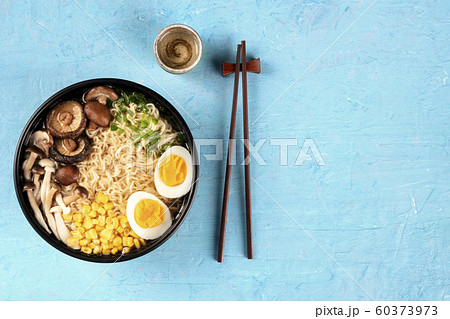 Ramen. Soba noodles with an egg, shiitake and enoki mushrooms, corn and scallions, overhead shot on a blue background with chopsticks, sake, and a place for text 60373973