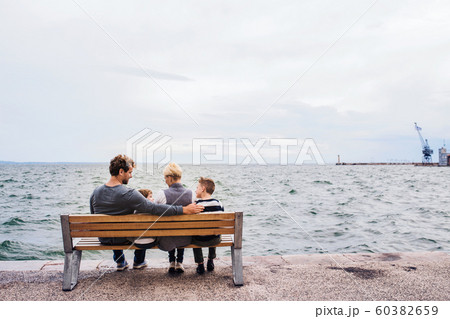 Rear view of young family with two small children on bench outdoors on beach. 60382659
