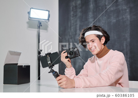 Young smiling male vlogger with photocamera sitting by desk and shooting himself Young smiling male vlogger with photocamera sitting by desk and shooting himself 60386105