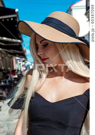 High fashion portrait of a graceful woman in an elegant straw hat and dress that walks along a city street 60387099