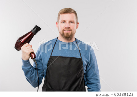 Young bearded hairdresser in apron and blue pullover holding hairdryer Young bearded hairdresser in apron and blue pullover holding hairdryer 60389123