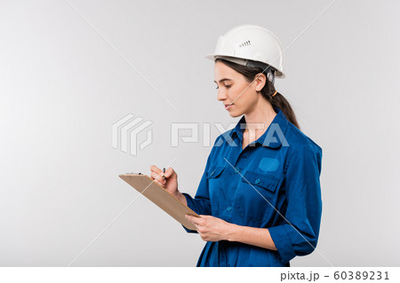 Young successful female engineer in blue workwear and safety helmet making notes Young successful female engineer in blue workwear and safety helmet making notes 60389231