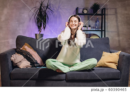 A young woman sitting on a sofa in a cozy room listening to music on wireless headphones. Girl happy. 60390465
