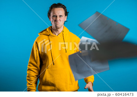 Serious man throwing papers on blue background. He is unsatisfied, anxious and confused with work. Young male student checks documents, reports. Serious man throwing papers on blue background. He is unsatisfied, anxious and confused with work. Young male student checks documents, reports. 60416473