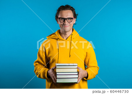 European student in yellow on blue background in studio holds stack of university books from library. Guy smiles, he is happy to graduate. European student in yellow on blue background in studio holds stack of university books from library. Guy smiles, he is happy to graduate. 60446730