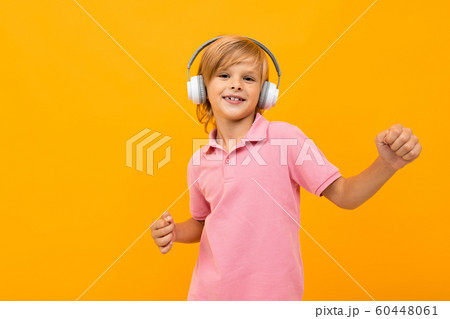 European blond boy in a pink T-shirt listens to music in white headphones on an orange background European blond boy in a pink T-shirt listens to music in white headphones on an orange background 60448061