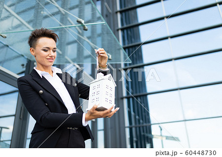 Caucasian woman in office clothes holds a little white house and waits colleague near the office Caucasian woman in office clothes holds a little white house and waits colleague near the office 60448730