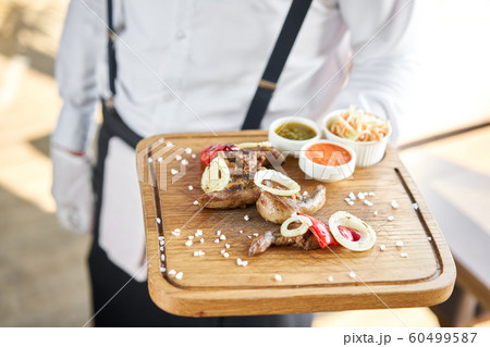 The waiter is holding a plate of Lamb tongue kebab with Pickled cabbage and two sauces. Serving on a wooden Board. Barbecue restaurant menu, a series of photos of different meats. The waiter is holding a plate of Lamb tongue kebab with Pickled cabbage and two sauces. Serving on a wooden Board. Barbecue restaurant menu, a series of photos of different meats. 60499587