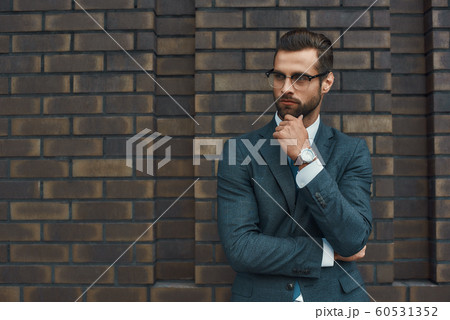 Lost in thoughts. Portrait of handsome bearded businessman in eyeglasses and formal wear thinking about something while standing against brick wall 60531352
