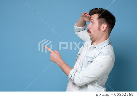 Young man pointing, surprised by something or someone, isolated on blue background. 60562569