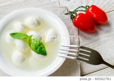 Mini mozzarella cheese balls with basil in a bowl and two cherry tomatoes on a rustic wooden table. Mini mozzarella cheese balls with basil in a bowl and two cherry tomatoes on a rustic wooden table. 60562761