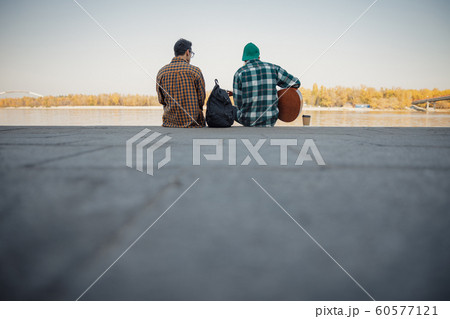 Two male friends siting with guitar near river 60577121