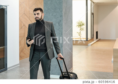 Young elegant businessman pulling suitcase while moving along hotel lounge 60596416