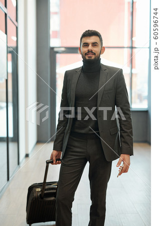 Young businessman with suitcase and smartphone standing inside airport Young businessman with suitcase and smartphone standing inside airport 60596744