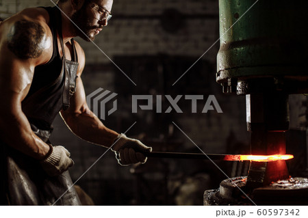 Young caucasian blacksmit in hot temperature while heating steel piece in furnace Young caucasian blacksmit in hot temperature while heating steel piece in furnace 60597342