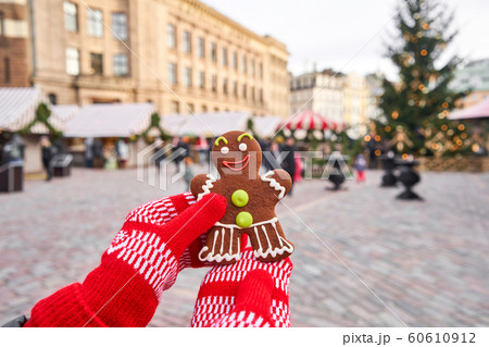 Hand in red mitten holding a smiling gingerbread man and christmas mood in blurred background. Christmas market in old town European small city. 60610912