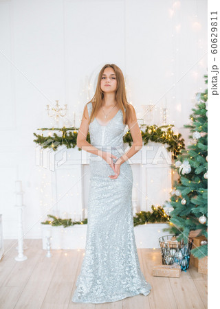 Attractive young girl with blond hair and evening make-up in an evening dress in a floor dress near a Christmas tree and a fireplace in a bright studio. New Year 2020 60629811