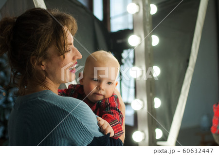 Attractive curly young mother in blue sweater hugging her toddler baby in red shirt in the room with Christmas decoration, Xmas tree and lights Attractive curly young mother in blue sweater hugging her toddler baby in red shirt in the room with Christmas decoration, Xmas tree and lights 60632447