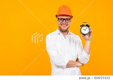 Beautiful caucasian engineer man with glasses and orange helmet holds an alarm clock isolated on Beautiful caucasian engineer man with glasses and orange helmet holds an alarm clock isolated on 60655432
