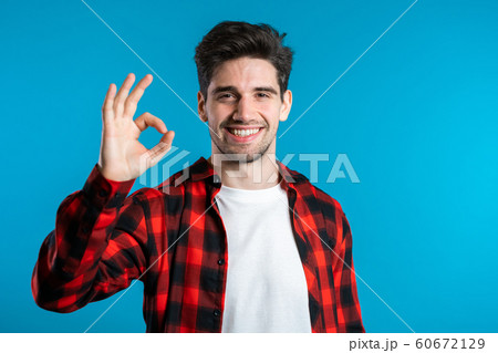 Handsome young man in red plaid shirt smiles to camera. Hipster guy showing OK sign over blue background. Winner. Success. Body language. Handsome young man in red plaid shirt smiles to camera. Hipster guy showing OK sign over blue background. Winner. Success. Body language. 60672129