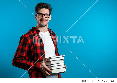 European student in red plaid shirt on blue background in studio holds stack of university books from library. Guy smiles, he is happy to graduate. European student in red plaid shirt on blue background in studio holds stack of university books from library. Guy smiles, he is happy to graduate. 60672198