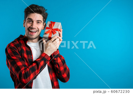Handsome man holding gift box on blue studio background smiles to camera. Happy european guy received present and interested in what's inside. Handsome man holding gift box on blue studio background smiles to camera. Happy european guy received present and interested in what's inside. 60672230