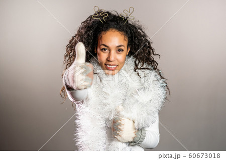 Young african american woman in knitted mittens warm sweater and deer horns smiling and showing thumbs up sign on gray studio background. Girl in good winter christmas mood. 60673018