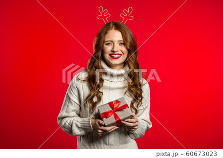 Young pretty woman smiling and holding gift box on red studio background. Girl with beautiful hairstyle in white sweater and deer horns. Christmas mood. 60673023