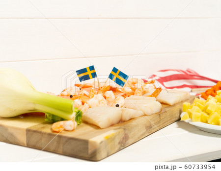 Salmon, cod, shrimps, fennel on a wooden cutting board. Potatoes and carrots in white plates. Swedish flags. Ingredients for traditional Scandinavian fish soup. 60733954