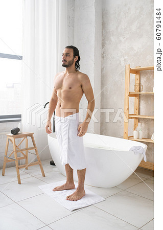 Young shirtless man with soft white towel on hips standing on small rug Young shirtless man with soft white towel on hips standing on small rug 60791484
