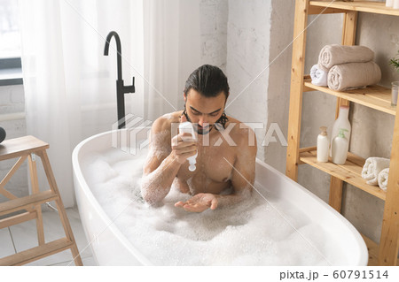 Wet young shirtless man using shower gel while having bath with foam 60791514