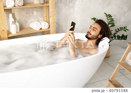 Young naked man watching video or scrolling in smartphone while lying in water Young naked man watching video or scrolling in smartphone while lying in water 60791588