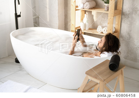 Young restful man scrolling in smartphone while taking bath with foam 60806458