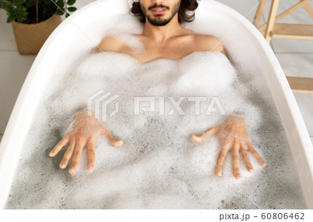 Young man lying in white bathtub filled with hot water and touching foam 60806462
