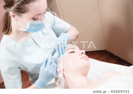 Close-up Attractive young woman gets anti-aging face injections. She lies calmly in a clinic or salon. An experienced young cosmetologist fills female wrinkles with hyaluronic acid from a syringe 60809591