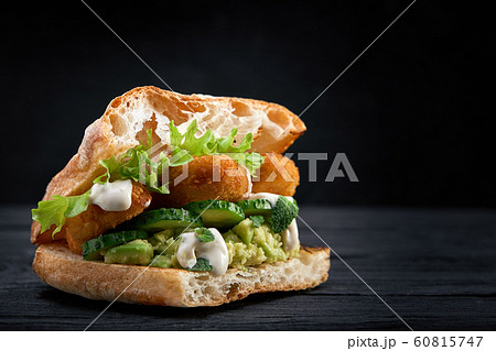 Appetizing sandwich on a wooden board. Baguette sandwich with filling from lettuce, slices tomato. Dark wooden background. View from above. Close-up. Macro photography. Appetizing sandwich on a wooden board. Baguette sandwich with filling from lettuce, slices tomato. Dark wooden background. View from above. Close-up. Macro photography. 60815747