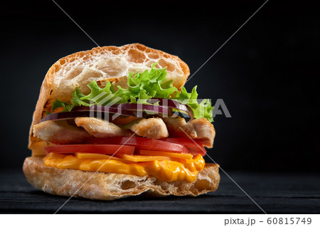 Appetizing sandwich on a wooden board. Baguette sandwich with filling from lettuce, slices tomato. Dark wooden background. View from above. Close-up. Macro photography. 60815749