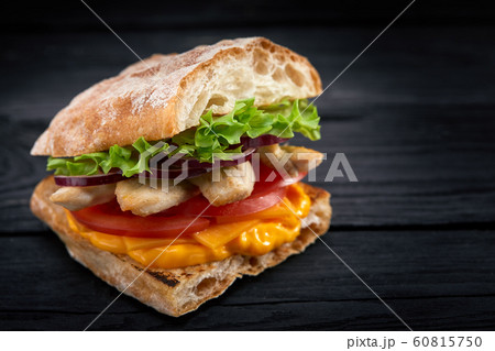 Appetizing sandwich on a wooden board. Baguette sandwich with filling from lettuce, slices tomato. Dark wooden background. View from above. Close-up. Macro photography. 60815750