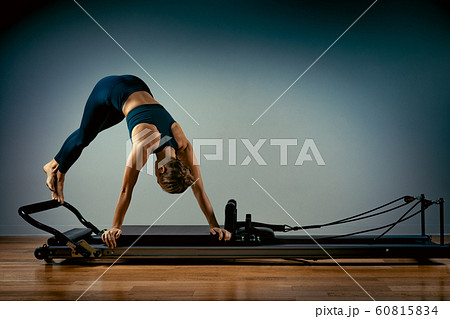 Young girl doing pilates exercises with a reformer bed. Beautiful slim fitness trainer on reformer gray background, low key, art light. Fitness concept Young girl doing pilates exercises with a reformer bed. Beautiful slim fitness trainer on reformer gray background, low key, art light. Fitness concept 60815834
