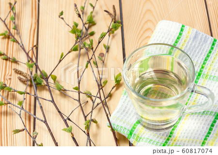 Birch juice on kitchen towel on a wooden table in a glass mug, next to a branch of birch with young leaves 60817074