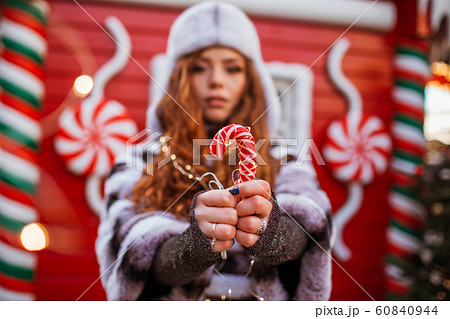 Outdoor portrait of young beautiful redhead happy smiling girl is wearing hat with traditional xmas lollipop. Festive red Christmas house on background. 60840944