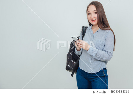 Young lovely asian student standing, holding a phone, smiling to the camera, backpack slung over her shoulder. Young lovely asian student standing, holding a phone, smiling to the camera, backpack slung over her shoulder. 60843815