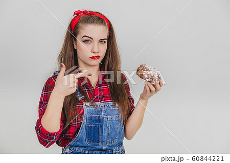 Nice young girl holding a big chocolate doughnut, pointing her finger at it. Judging by her face expression she doesn't like such sweeties. Nice young girl holding a big chocolate doughnut, pointing her finger at it. Judging by her face expression she doesn't like such sweeties. 60844221