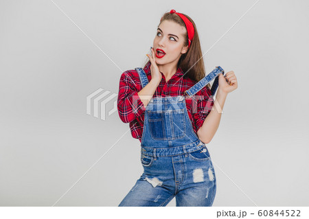 Beautiful young girl in red standing, keeping one hand on her cheek, posing, pulling up her suspender. Surprised look. Beautiful young girl in red standing, keeping one hand on her cheek, posing, pulling up her suspender. Surprised look. 60844522