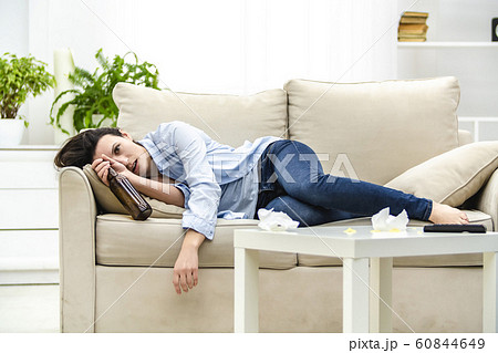 Young drunk woman with empty bottle in her hand is lying on the sofa. Woman feels headache. 60844649