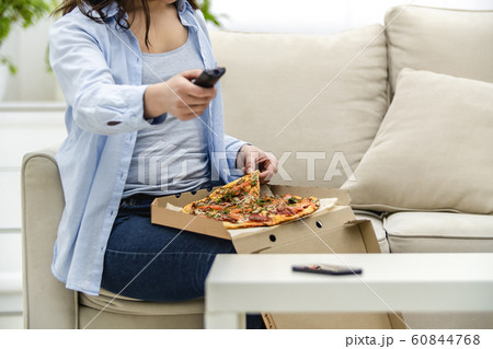 Crop. Unrecognizable woman is sitting on her sofa at home and is watching television while eating pizza. 60844768