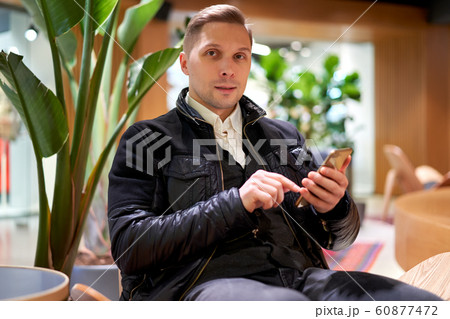 Male in leather jacket with smartphone looking at camera while sitting on chair in store against green plant 60877472