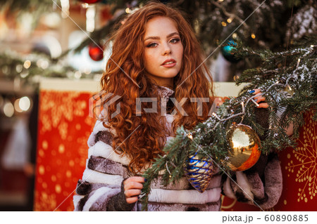 Outdoor portrait of young beautiful redhead happy smiling girl is wearing fur coat near decorated Christmas tree. 60890885