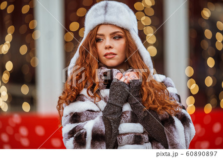 Outdoor portrait of young beautiful redhead happy smiling girl is wearing fur warm hat. Festive Christmas lights on background. Outdoor portrait of young beautiful redhead happy smiling girl is wearing fur warm hat. Festive Christmas lights on background. 60890897