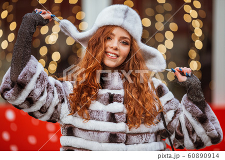 Outdoor portrait of young beautiful redhead happy smiling girl is wearing fur warm hat. Festive Christmas lights on background. 60890914